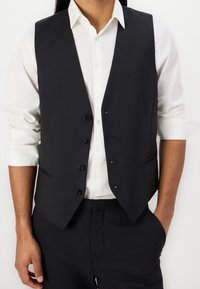 Schwarze Weste aus glattem Stoff, mit einem V-Ausschnitt, sechs Knöpfen und zwei seitlichen Taschen. Wird über einem weißen Hemd getragen.