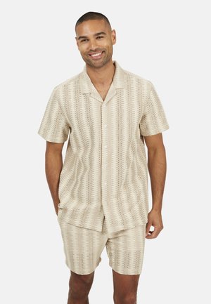 Lachende man in een beige overhemd met korte mouwen en patroon, gecombineerd met bijpassende korte broek, staand tegen een witte achtergrond.