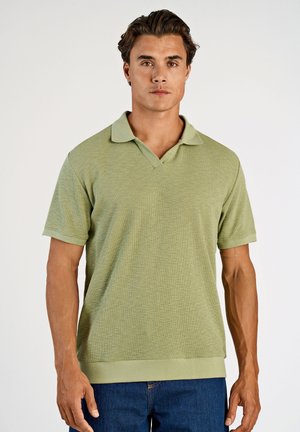 Jonge man met kort donker haar, gekleed in een lichtgroen overhemd met korte mouwen en een kraag, en een blauwe spijkerbroek, staand tegen een effen achtergrond.