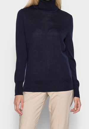 Femme portant un pull à col roulé bleu marine foncé associé à un pantalon beige clair, debout devant un fond clair uni.