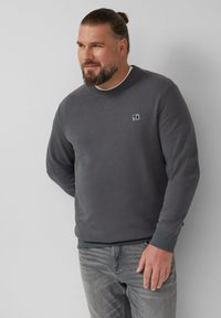 Uomo con barba e capelli legati, che indossa una felpa grigio antracite e jeans grigi sbiaditi, in piedi contro un muro uniforme di grigio chiaro.