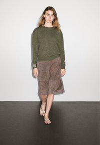 Maglione a maglia verde oliva, gonna in suede marrone e infradito argentati. La gonna ha una forma fluida con un orlo che arriva sotto il ginocchio.