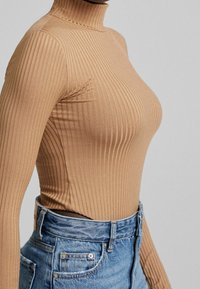 Kvinna i en figurnära, ribbad kamelturtleneck som är instoppad i högmidjade blå denimjeans mot en enkel grå bakgrund.