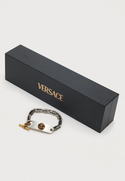 Μαύρο κουτί με χρυσή επιγραφή "Versace" δίπλα σε ασημένιο και χρυσό βραχιόλι αλυσίδα με χρυσό έμβλημα Μέδουσα και κούμπωμα τύπου toggle.