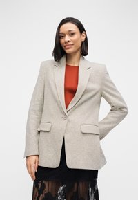 Lichtgrijs colbert met een gestructureerde afwerking, enkele knoopsluiting en twee voorzakken, gedragen over een rood topje en gecombineerd met een zwarte rok.