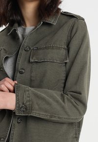 Dżinsowa kurtka w oliwkowej zieleni z guzikami, dwoma kieszeniami na piersi i luźnym krojem. Posiada klasyczny kołnierz oraz lekko fakturowaną powierzchnię.