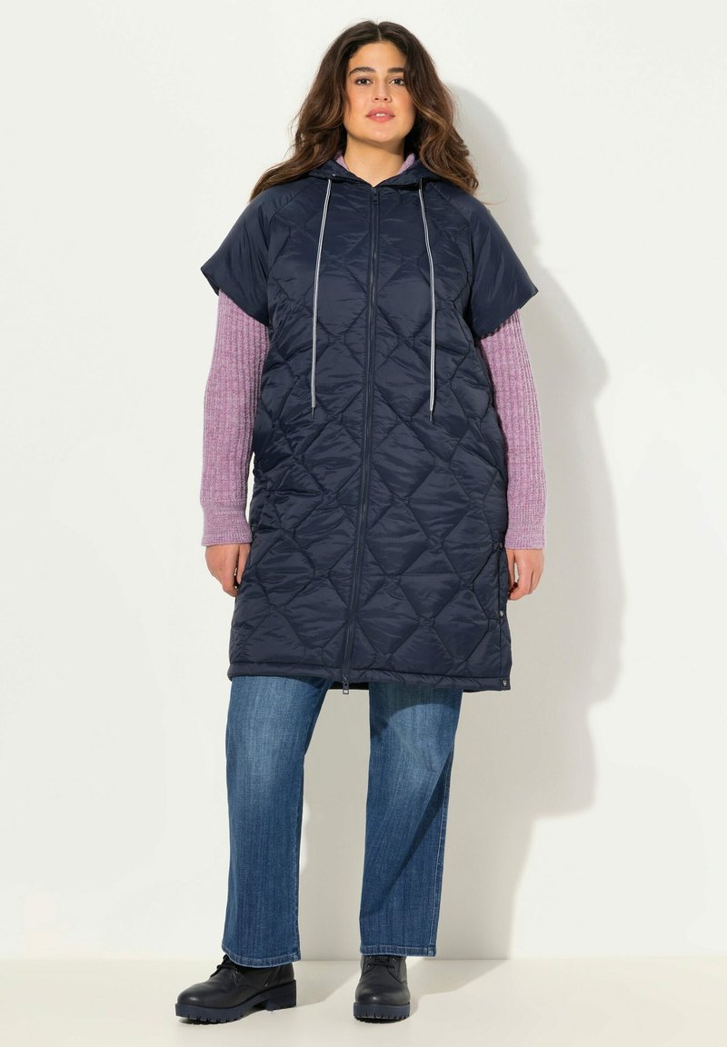 Námořnický prošívaný dlouhý vest s kapucí, krátkými rukávy, předním zipem a bílými šňůrkami, přes růžový top s dlouhými rukávy a džíny.