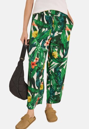 Osoba nosząca luźne zielone spodnie w tropikalny wzór, brązowe buty typu slip-on, trzymająca czarną pikowaną torbę i mająca na sobie biały top.