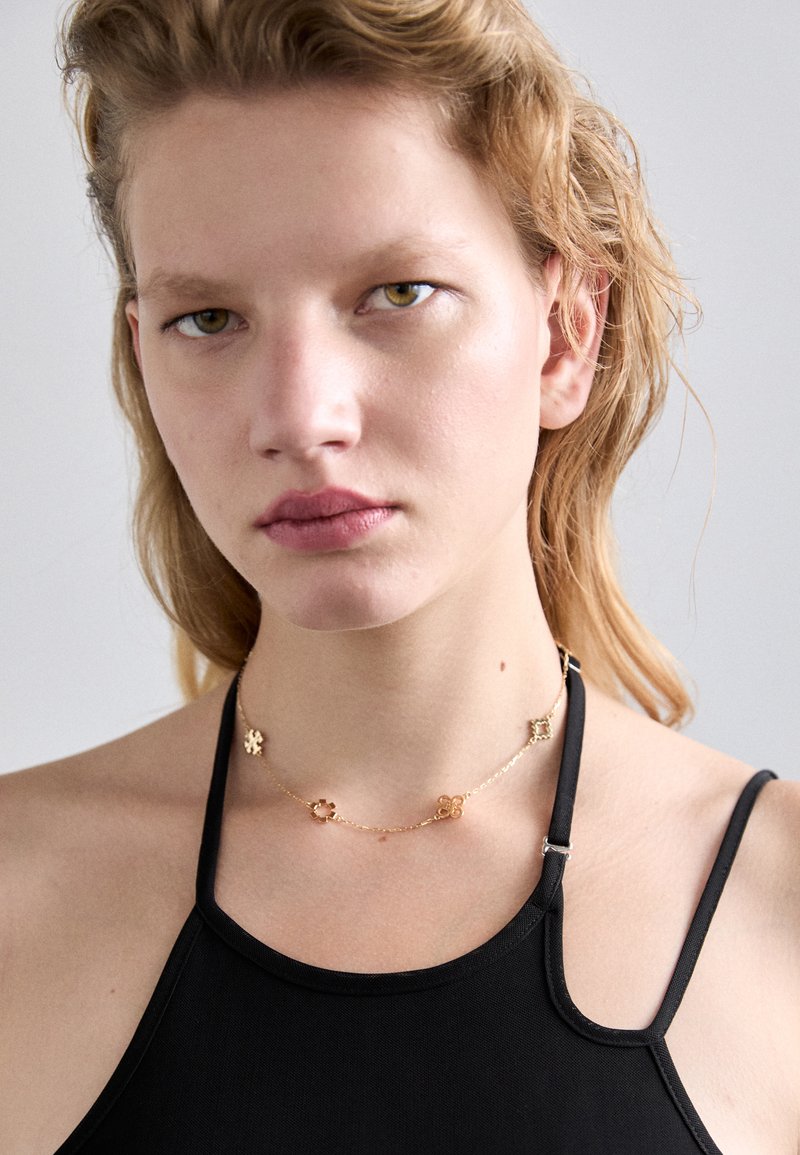 Gold chain necklace featuring decorative metal floral shapes and accents, worn over a black tank top, highlighting its design and texture.