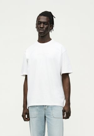 Uomo con pelle scura e dreadlocks indossa una semplice maglietta bianca oversize e jeans azzurri, in piedi davanti a uno sfondo neutro.