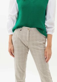 Veste en tricot vert portée sur un chemisier blanc, associée à un pantalon à carreaux clair avec fermeture à boutons et texture subtile.