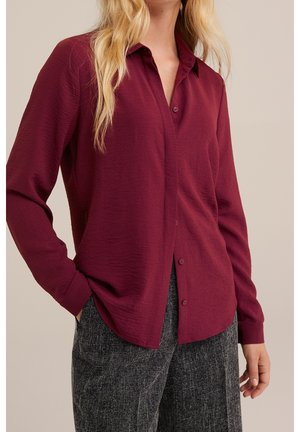 Persona bionda che indossa una camicia bordeaux a maniche lunghe con bottoni e pantaloni grigi strutturati, in piedi con una mano in tasca davanti a uno sfondo neutro.