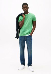 Zielona bawełniana koszulka z białym kołnierzykiem, połączona z niebieskimi dżinsami. Model nosi białe trampki, trzyma granatową kurtkę na ramieniu.