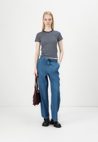 Striped short-sleeve shirt in navy and white. Blue wide-leg trousers with front tie. Model holds a burgundy bag and wears black shoes.