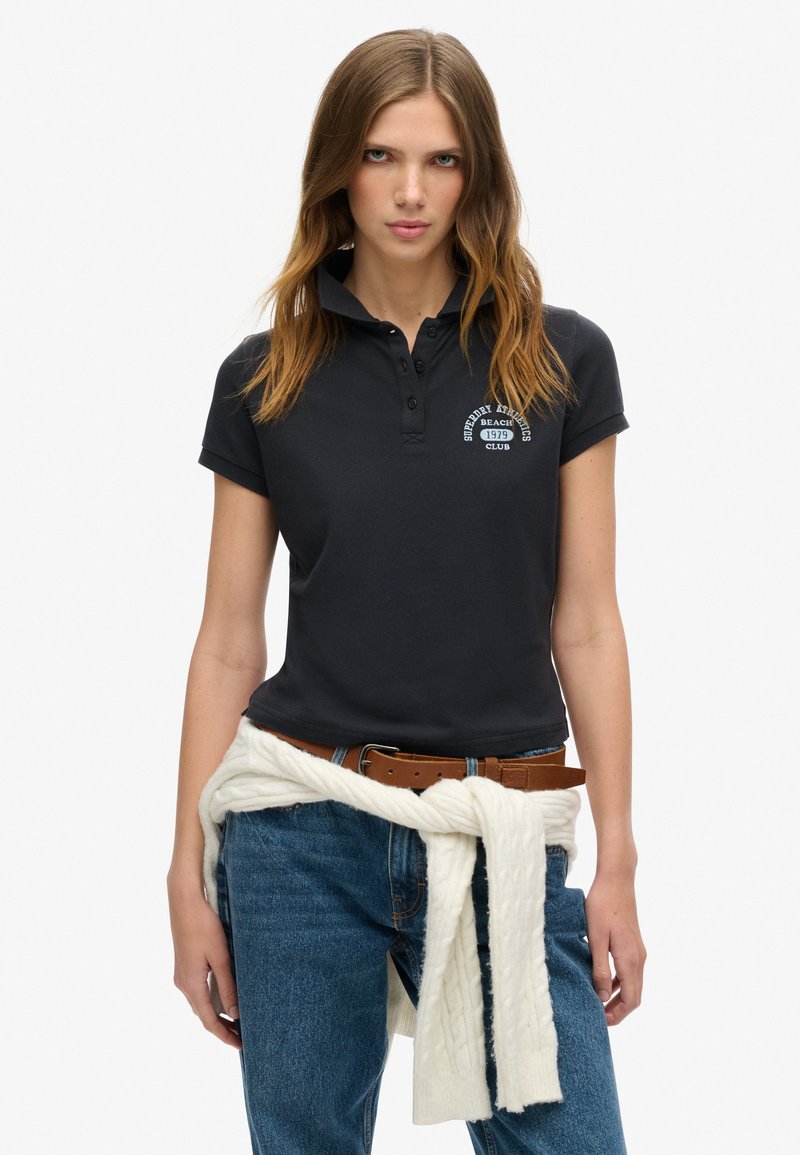 Polo de color azul marino oscuro con mangas cortas y cuello. Presenta un bordado blanco de un logo. Se lleva con jeans azules y un suéter de punto color crema atado a la cintura.