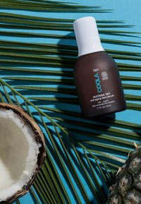 Braune Flasche mit weißem Deckel, beschriftet mit "Coola Sunless Tan Anti-Aging Face Serum", platziert zwischen grünen Palmblättern und einer Kokosnussschale auf blauem Hintergrund.