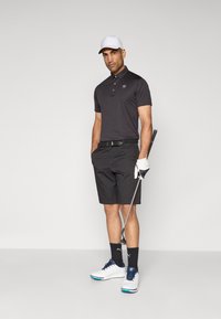 Čierna polokošeľa a šortky, biele golfové topánky s modrými akcentmi, biela čiapka, drží golfovú palicu, má na ruke bielu rukavicu, neutrálny pozadie.