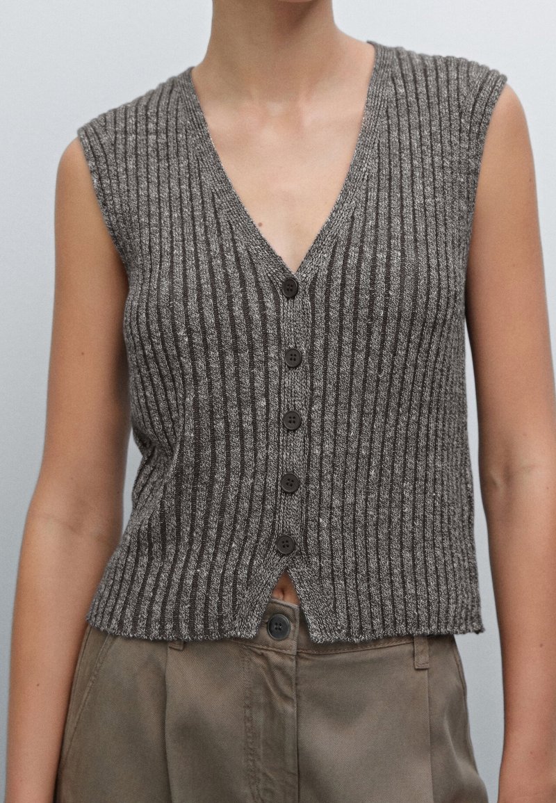 Gilet gris en maille côtelée sans manches avec des boutons noirs porté sur un pantalon beige taille haute, montré des épaules aux hanches.