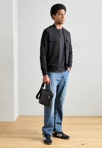 Jachetă bomber neagră cu buzunare, tricou negru, blugi decolorati bleumarin, geantă neagră de umăr și adidași negri cu dungi albe.