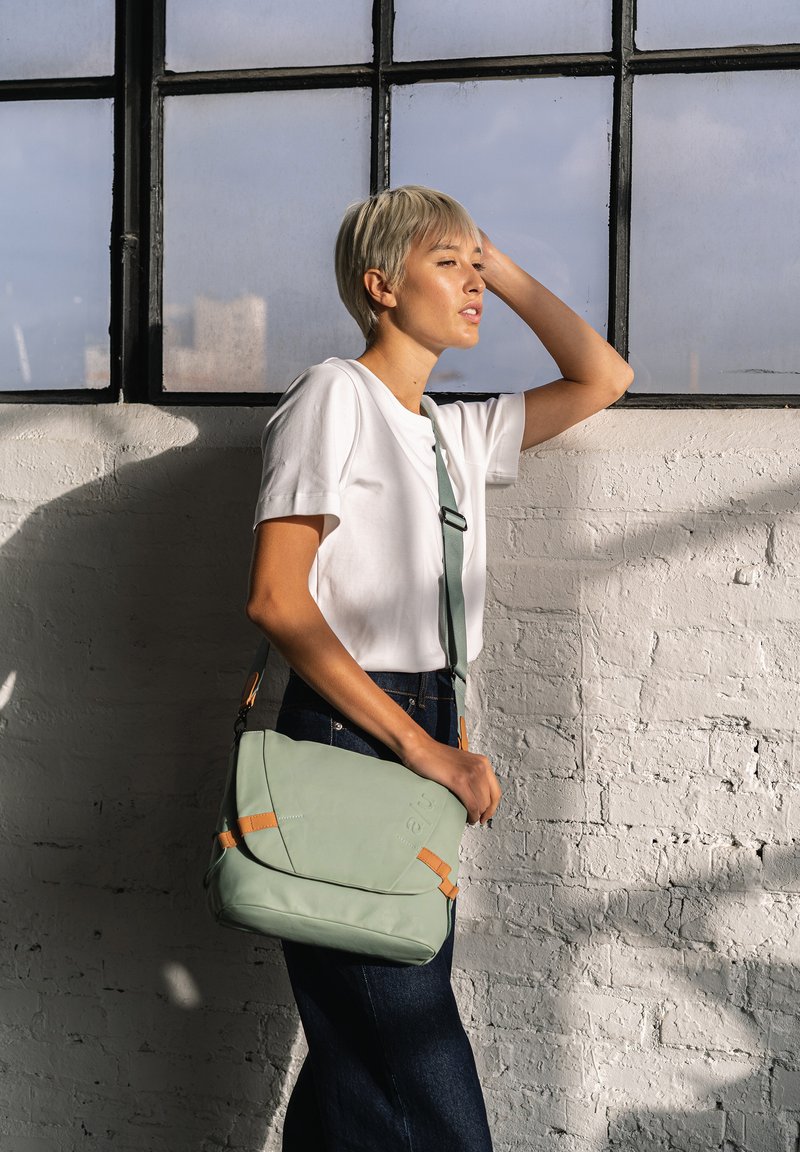 Hellgrünes Leder-Crossbody-Täschchen mit orangefarbenen Akzenten, umklappbarem Deckel und verstellbarem Trageriemen. Das Modell trägt ein weißes T-Shirt und Jeans.