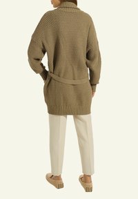 Frau, die mit den Händen in den Taschen steht und eine lockere braune Strickjacke, beige Hosen und passende braune Slippers trägt, von hinten betrachtet.