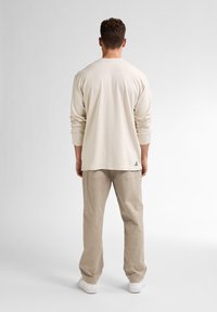 Långärmad beige t-shirt med avslappnad passform och rund halsringning, matchat med ljusbeiga byxor och vita sneakers. Diskret logotypdetalj vid fållen.