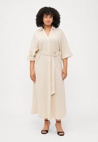A woman with curly hair wears a beige belted dress and black heeled sandals, standing against a plain white background.