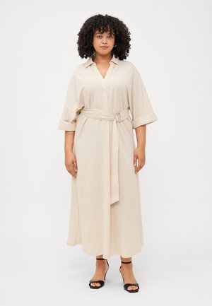 A woman with curly hair wears a beige belted dress and black heeled sandals, standing against a plain white background.