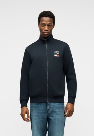 Mann trägt eine schwarze Zip-up-Jacke mit Tommy-Hilfiger-Logo auf der Brust und blaue Jeans, steht vor einem einfarbigen hellen Hintergrund.