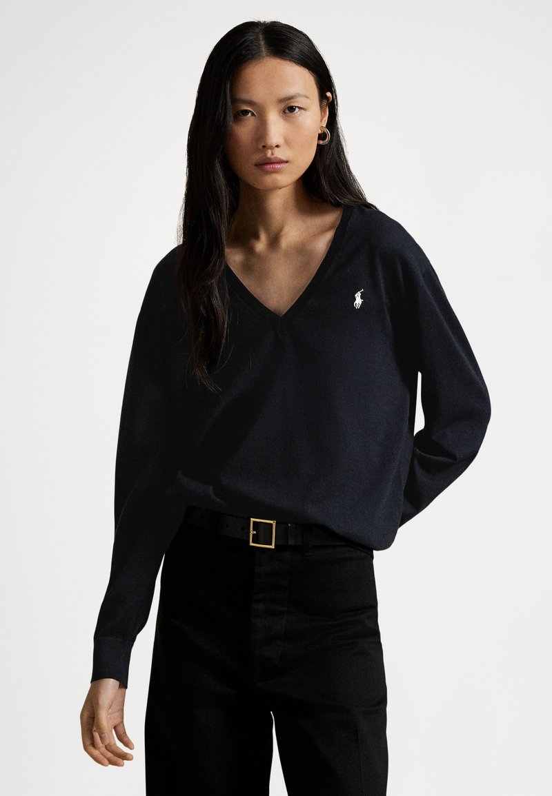 Black V-neck long-sleeve top made of soft fabric with a small white logo. Worn with high-waisted black trousers and a gold-buckled belt.
