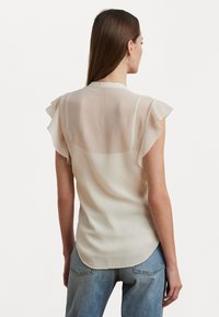 Sheer cream blouse with ruffled cap sleeves, a round neck, and a curved hem, paired with light blue denim jeans.