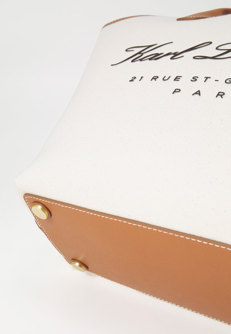 Sac fourre-tout en toile avec des accents en cuir marron, présentant des rivets en laiton et un texte signature en noir. Le design est structuré, avec une texture lisse.
