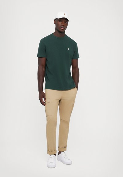 T-shirt en coton vert avec un petit logo, associé à un pantalon cargo beige avec des poches, et des sneakers blancs, complété par une casquette blanche.