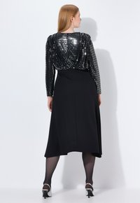 Černé šaty s vrškem zdobeným flitry a dlouhými rukávy, s volánovou sukní a přiléhavým pasem, doplněné vysokými podpatky.