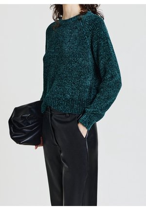Femme portant un pull en maille texturée bleu sarcelle et un pantalon en cuir noir, tenant une grande pochette en cuir noir dans une main.