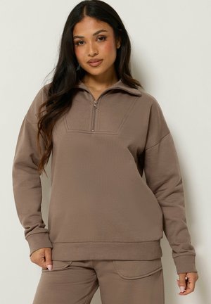 Kvinde iført en taupe lynlåstrøje og matchende bukser, stående mod en neutral baggrund med afslappet kropsholdning.