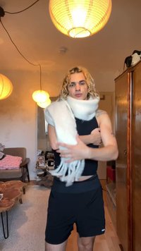 Écharpe blanche et duveteuse enroulée autour des épaules, avec des franges aux extrémités. Portée sur un haut noir ajusté et un short de sport. Cadre intérieur chaleureux.