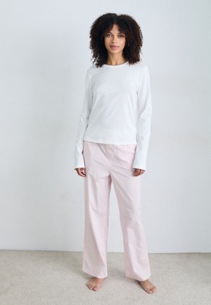 Une femme se tient pieds nus dans une pièce lumineuse, portant une chemise blanche à manches longues et un pantalon large rose pâle, avec des cheveux bouclés entourant son visage.