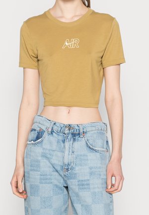 Personne portant un crop top moutarde avec le logo "AIR" blanc et un jean en denim à carreaux bleu clair taille haute, sur fond blanc.