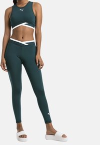 Turkusowy stanik sportowy i legginsy z białymi akcentami, wykonane z elastycznego materiału. Model nosi białe klapki bez pięty.