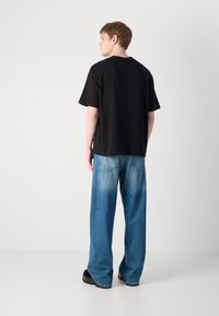 Svart bomullst-skjorte med korte ermer, kombinert med blå vide denimjeans med falmede seksjoner og baklommer, iført svarte sko.