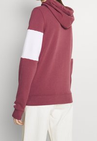 Persona con una sudadera con capucha color granate, con una franja blanca en la parte superior del brazo y pantalones blancos, vista desde atrás.