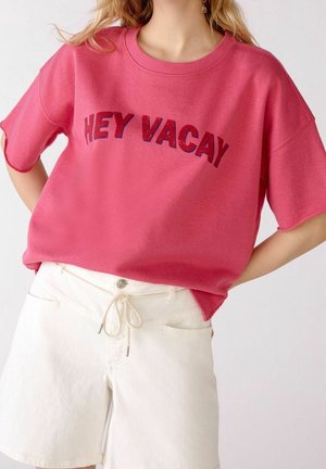 Vrouw die een roze T-shirt met korte mouwen draagt met de tekst "HEY VACAY" en witte shorts met trekkoord, staand met de handen in de zij.