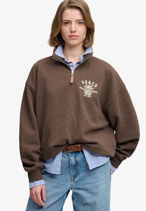 Junge Person trägt einen braunen Sweatshirt mit Viertelreißverschluss über einem blau-rosa gestreiften Hemd mit Knöpfen und hellblauen Jeans mit geflochtenem Gürtel.