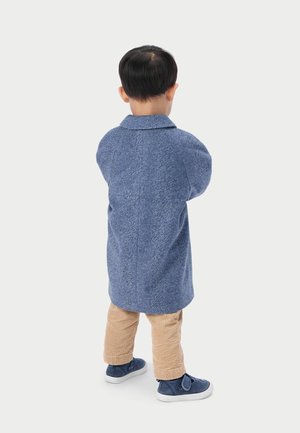 Enfant debout, vu de dos, portant un manteau bleu, un pantalon en velours côtelé beige et des baskets bleu marine sur un fond blanc uni.