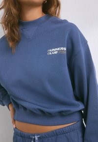 Felpa blu corta con colletto e polsini a coste, dotata della scritta "RUNNERS CLUB" stampata in bianco e oro. Tessuto testurizzato, vestibilità comoda.