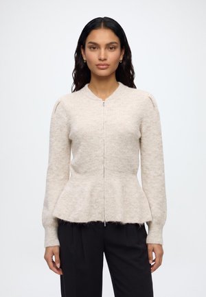 Femme aux longs cheveux foncés portant un pull péplum zippé beige clair et un pantalon noir, debout devant un fond uni.
