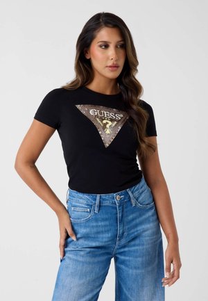 Femme aux cheveux longs et ondulés portant un t-shirt noir Guess avec un logo triangulaire marron et un jean bleu, debout avec une main sur la hanche.