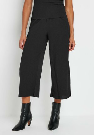 Kvinde iført sorte vidde-bukser med korte ben og sorte ankelstøvler med hæl mod en ensfarvet baggrund.