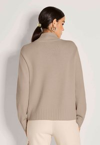 Frau mit dunklem Haar in einem tiefen Pferdeschwanz, die goldene Creolenohrringe und einen beigen, gerippten Pullover trägt, kombiniert mit hellen Hosen, von hinten gesehen.